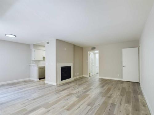 Apartments  for rent in Encino A vacant room with light wood flooring, neutral-colored walls, a fireplace in the corner, an open doorway, and a kitchen area visible in the background. White Oak Apartments in Encino 5465 White Oak Avenue Encino, CA 91316  P: 866-471-5691 TTY: 711 F: 818-647-0393